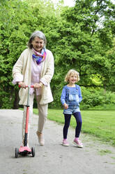 Active senior woman riding push scooter while granddaughter walking in park - ECPF00943