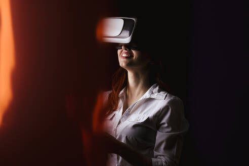 Junge Frau lächelt, während sie eine VR-Brille auf schwarzem Hintergrund benutzt - LJF01535