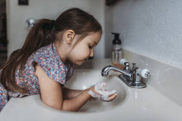 Side view of young girl washing hands in sink with soap - CAVF83491