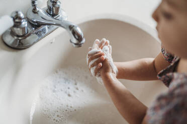 High angle view of young girl washing hands in sink with soap - CAVF83493