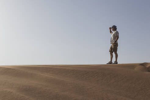 Male tourist shielding eyes while standing on sand dune against clear sky at Dubai, United Arab Emirates - SNF00247