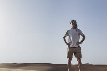 Lächelnder Tourist, der mit den Händen auf der Hüfte vor dem klaren Himmel in der Wüste in Dubai, Vereinigte Arabische Emirate, steht - SNF00248