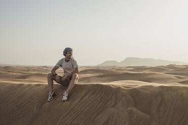 Männlicher Tourist schaut weg, während er auf Sanddünen in der Wüste von Dubai, Vereinigte Arabische Emirate, sitzt - SNF00256