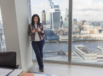 Businesswoman texting with smart phone at urban office window - CAIF27537