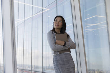 Nachdenkliche Geschäftsfrau mit digitalem Tablet am Fenster eines Hochhauses - CAIF27540