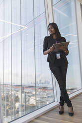 Selbstbewusste Geschäftsfrau mit digitalem Tablet am Fenster eines Hochhausbüros - CAIF27585