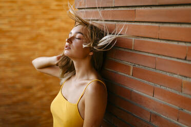 Beautiful young woman with tousled hair standing against brick wall in balcony - TCEF00716