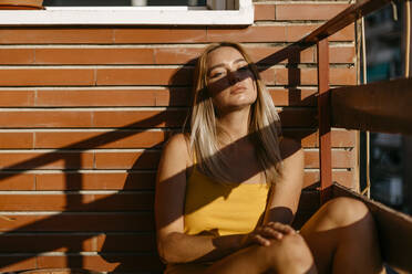 Sunlight on young woman relaxing while sitting against brick wall in balcony - TCEF00719