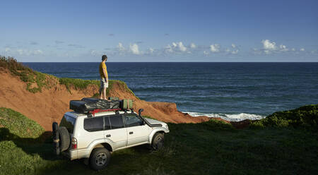 Männlicher Tourist steht auf dem Dach eines 4x4 und schaut auf das Meer vor blauem Himmel - VEGF02316