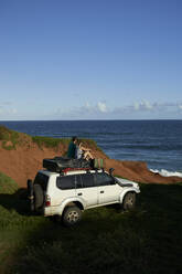 Mid adult couple sitting on 4x4 roof while looking at sea against blue sky - VEGF02322