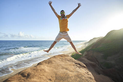 Fröhlicher Mann, der auf eine Felsformation am Strand springt, gegen blauen Himmel, lizenzfreies Stockfoto
