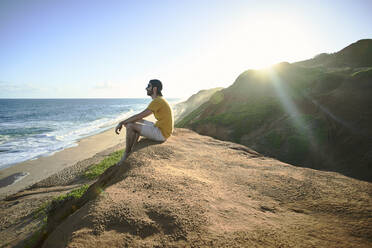 Mann sitzt auf einer Felsformation und betrachtet das Meer vor dem Himmel - VEGF02329