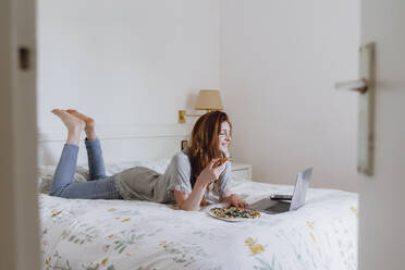 Frau isst Pizza, während sie im Schlafzimmer einen Film auf dem Laptop schaut - AFVF06410
