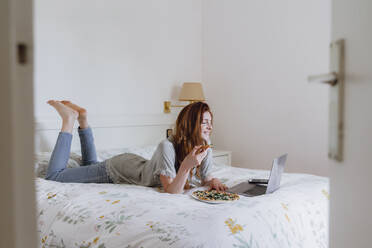 Woman eating pizza while watching movie on laptop in bedroom - AFVF06410