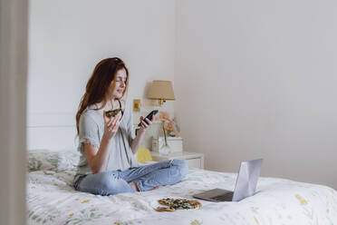 Beautiful woman using mobile phone while sitting with pizza and laptop on bed at home - AFVF06412