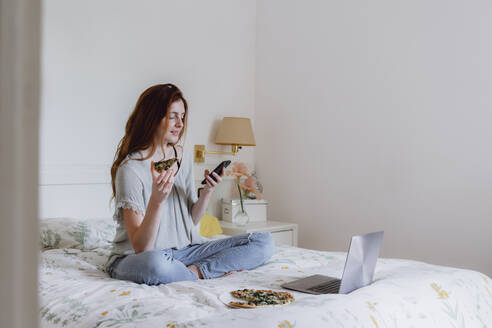 Schöne Frau, die ein Mobiltelefon benutzt, während sie mit Pizza und Laptop auf dem Bett zu Hause sitzt - AFVF06412