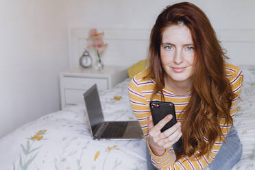 Schöne Frau mit grauen Augen und langen braunen Haaren sitzt mit Handy auf Schlafzimmer zu Hause - AFVF06415