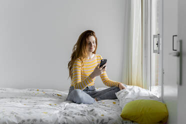 Lächelnde junge Frau, die ein Smartphone benutzt, während sie zu Hause auf dem Bett am Fenster sitzt - AFVF06416