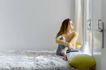 Nachdenkliche Frau schaut durch das Fenster, während sie zu Hause auf dem Bett sitzt - AFVF06417