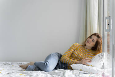 Thoughtful smiling woman while lying on bed by window at home - AFVF06420