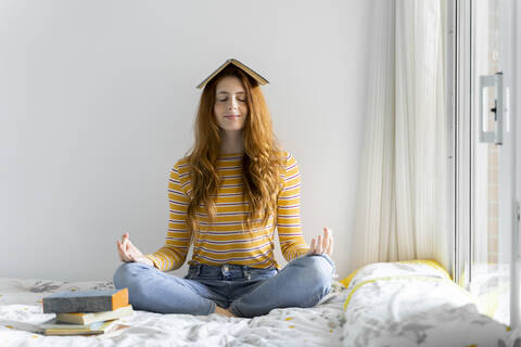 Young woman meditating in lotus position with book on head at home stock photo