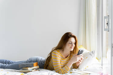 Smiling woman using mobile phone while lying on bed at home - AFVF06427