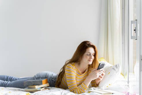 Lächelnde Frau, die ein Mobiltelefon benutzt, während sie zu Hause auf dem Bett liegt - AFVF06427