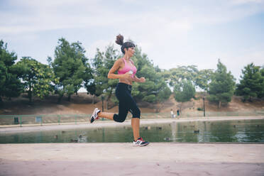 Dedicated active woman running while listening music by pond at park - OCMF01317