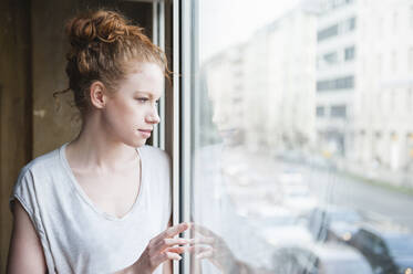 Nachdenkliche junge Frau, die durch das Fenster ihres Hauses schaut - DIGF12464