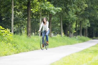 Smiling woman riding bicycle on footpath against green trees at park - DIGF12476