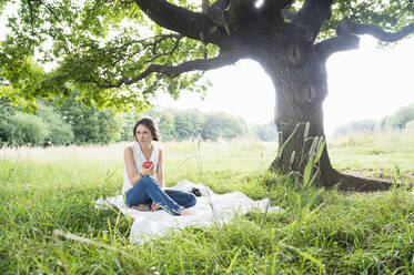 Nachdenkliche junge Frau schaut weg, während sie mit einem Apfel auf einer Picknickdecke unter einem Baum im Park sitzt - DIGF12482