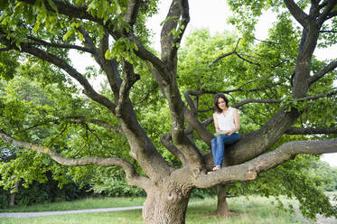 Young woman writing while sitting on tree branch at park - DIGF12483