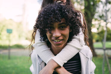 Nahaufnahme eines lächelnden Jungen mit lockigem Haar, der seine Schwester im Park huckepack nimmt - DCRF00189