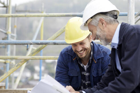 Happy architect and worker discussing building plan on scaffolding on a construction site - MJFKF00212