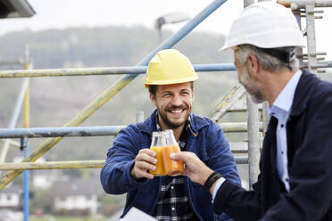 Glücklicher Architekt und Arbeiter auf einem Baugerüst auf einer Baustelle stoßen mit Saftflaschen an - MJFKF00214