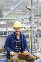 Portrait of a happy worker having a break on scaffolding on a construction site - MJFKF00217