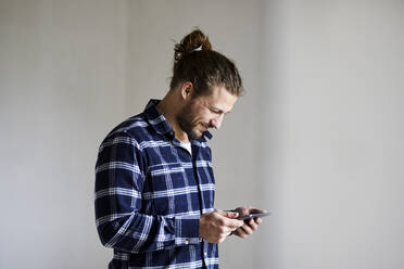 Lächelnder junger Mann mit kariertem Hemd und Smartphone - MJFKF00229