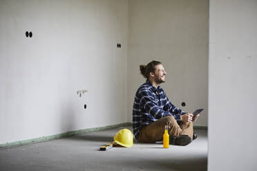 Glücklicher Arbeiter auf einer Baustelle, der auf dem Boden sitzend eine Pause macht - MJFKF00231