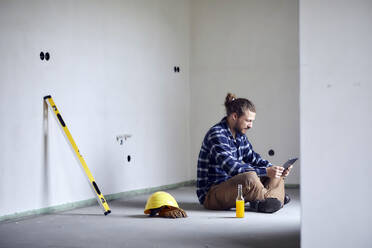 Arbeiter auf einer Baustelle, der auf dem Boden sitzt und ein Tablet benutzt - MJFKF00232