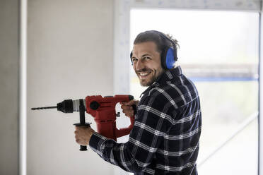 Porträt eines lächelnden Arbeiters, der eine elektrische Bohrmaschine auf einer Baustelle benutzt - MJFKF00234