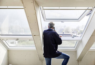 Mature man on the phone at the window on a construction site - MJFKF00273