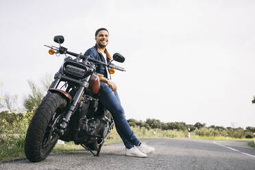 Smiling young biker leaning on motorcycle on road against sky - ABZF03156