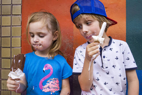 Niedliche Geschwister essen Eis am Stiel, während sie an der Wand stehen - JLOF00414