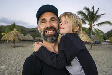 Bearded mature man carrying son while standing at beach - JLOF00417