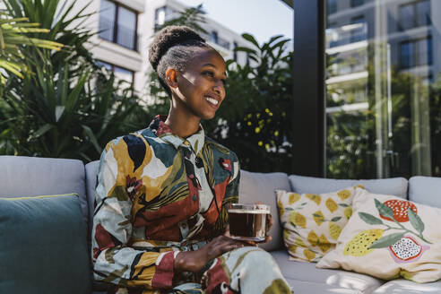 Porträt einer glücklichen jungen Frau, die auf einer Couch im Garten sitzt und schwarzen Kaffee trinkt - MFF05795