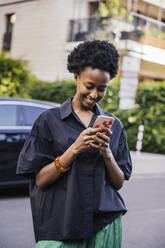 Portrait of smiling young woman standing on the street looking at cell phone - MFF05839