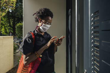 Junge Frau mit Textilschutzmaske steht vor der Eingangstür und benutzt ihr Smartphone - MFF05844