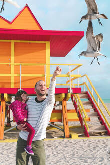 Glücklicher Vater, der seine Tochter trägt und Möwen füttert, während er am Strand von Miami, Florida, USA, steht - GEMF03799