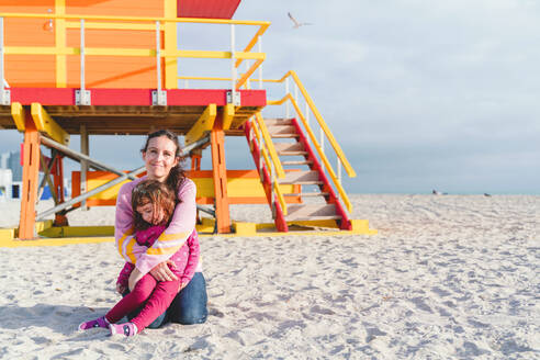 Loving mother embracing daughter at Miami beach against lifeguard hut, Florida, USA - GEMF03800