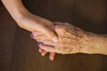 Close-up of granddaughter holding grandmother's hand on wooden table at home - DIGF12492